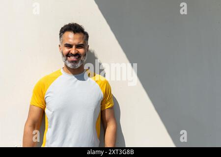 Allegro uomo indiano con una t-shirt gialla e bianca sorride brillantemente, guardando direttamente la telecamera sotto il sole estivo, gettando un'ombra forte su una Foto Stock