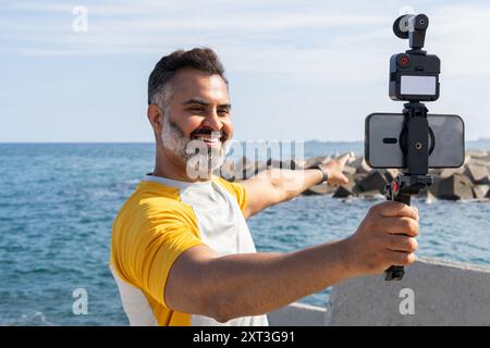 Un uomo indiano sorridente registra un vlog su uno smartphone montato su un gimbal, in piedi in riva al mare. Vestito con una t-shirt estiva, punta con orgoglio al Foto Stock