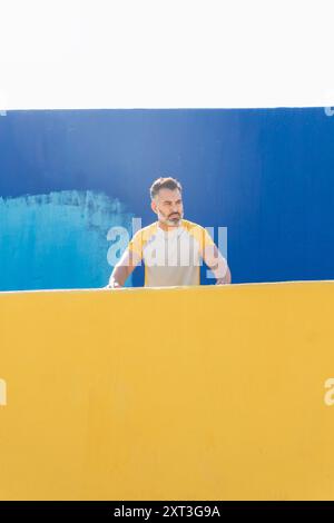 Un uomo indiano di abbigliamento sportivo giallo e bianco poggia su una parete gialla contro una parete blu a contrasto, guardando lontano dalla fotocamera durante una giornata estiva. Foto Stock