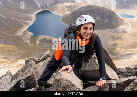 Una donna sorridente alza il terreno roccioso verso la cima di un vulcano dormiente, guardando la telecamera. E' vestita con un casco, uno zaino e. Foto Stock