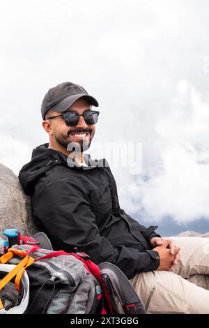 L'allegro escursionista maschile con occhiali da sole si prende una pausa su una roccia mentre cammina in montagna, guardando la macchina fotografica con un'espressione gioiosa. Foto Stock
