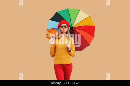 Vestito in stile autunnale, giovane donna elegante e allegra, con foglie d'acero gialle in posa con ombrello colorato che indossa un cappello rosso di berretto francese, k gialle Foto Stock