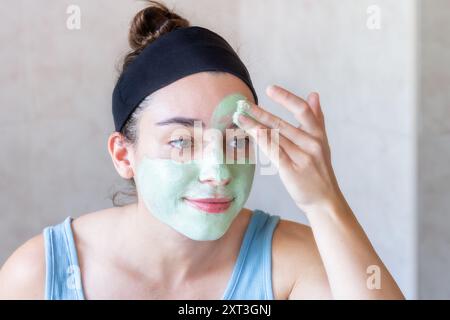 Una giovane donna che indossa un cerchietto applica una maschera facciale verde in un ambiente bagno, riflettendo un momento di cura personale e trattamento di bellezza Foto Stock