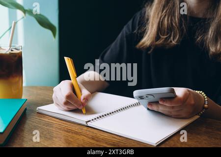 Vista ritagliata irriconoscibile delle mani di una studentessa che scrive su un notebook tenendo contemporaneamente in mano uno smartphone. Una rinfrescante bevanda ghiacciata si siede Foto Stock