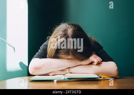 Studentessa corta irriconoscibile esausta, riposando la testa sulle braccia di fronte a un libro aperto, raffigurante il concetto di affaticamento scolastico Foto Stock