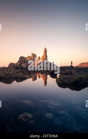 Spettacolari rocce marine che si riflettono su una tranquilla piscina di marea all'alba, con una figura solitaria che ammira la scena a Llanes, Asturie, con Playa de Pendueles Foto Stock