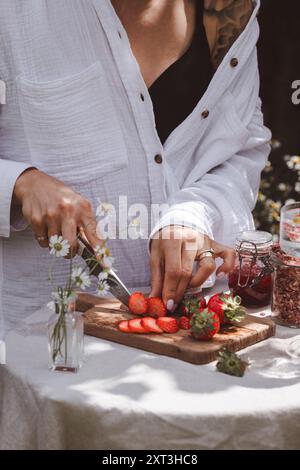 Donna corta e irriconoscibile che affetta fragole fresche su una tavola di legno per una sana colazione all'aperto, circondata da fiori e luce naturale. Foto Stock