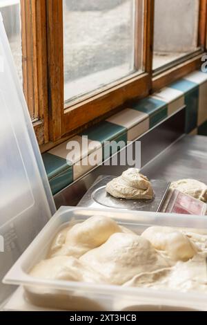 L'immagine cattura le palline di impasto fresco per pizza che si ripresentano su una bilancia e si appoggiano in un contenitore di plastica da una finestra soleggiata, mostrando i passaggi iniziali di pi Foto Stock