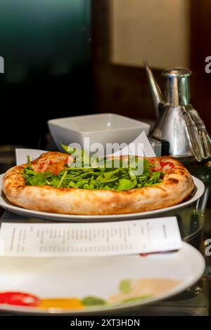 Una deliziosa pizza appena sfornata, condita generosamente con rucola fresca, servita su un piatto bianco in un elegante ristorante, accompagnata da un Met Foto Stock