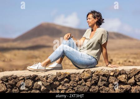 Una donna guarda con attenzione mentre è seduta su un muro di pietra, con un paesaggio arido sullo sfondo. Foto Stock