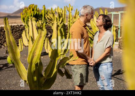 Un uomo e una donna maturi si tengono per mano e si guardano l'un l'altro, circondati da alte piante di cactus sotto un cielo limpido. Entrambi sono vestiti casualmente, l'uomo in una Foto Stock