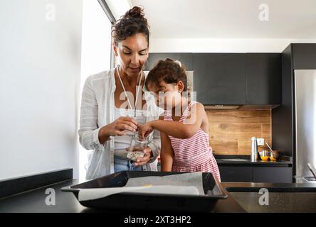 Una scena commovente in una cucina moderna dove una donna e sua figlia stanno preparando biscotti insieme, concentrandosi sull'aggiunta di sprinkles al doug Foto Stock