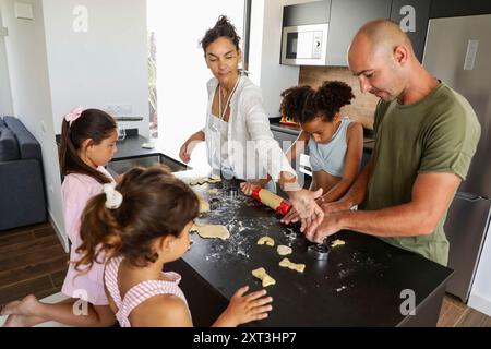 Una famiglia di cinque persone ama cucinare i biscotti, con tre bambini coinvolti nel processo di laminazione dell'impasto, guidati dai genitori in un elegante e contemporaneo ki Foto Stock