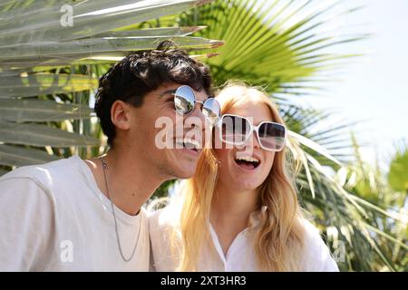 Una coppia gioiosa ride all'ombra delle palme, indossando eleganti occhiali da sole e crogiolandosi alla luce calda del sole Foto Stock