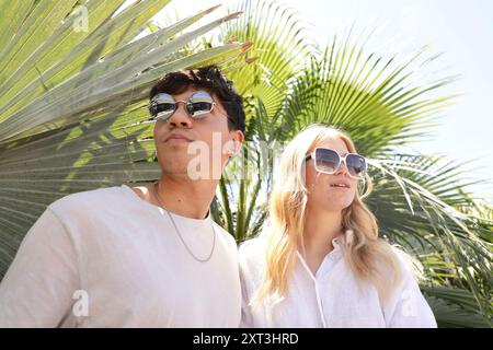 Una giovane coppia elegante con occhiali da sole si erge sotto le fronde delle palme, crogiolandosi alla luce del sole, trasudando eleganza informale e vibrazioni estive Foto Stock
