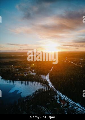 Uno scatto aereo cattura la bellezza di un tramonto in Lapponia, evidenziando un lago parzialmente ghiacciato e strade serpeggianti tra vasti tratti di foresta. Foto Stock