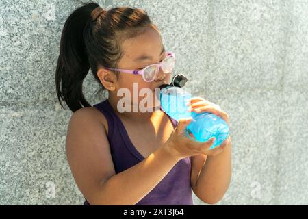 Una giovane ragazza con gli occhiali che indossa un top viola beve da una bottiglia d'acqua sportiva blu, appoggiata su una parete testurizzata grigia Foto Stock