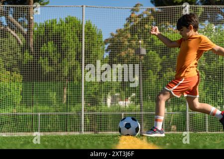 Un ragazzo che indossa una t-shirt arancione e pantaloncini corti gioca a calcio, calciando la palla su un lussureggiante campo verde mostrando energia ed entusiasmo Foto Stock