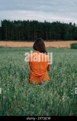 La vista posteriore di una donna bruna irriconoscibile, che guarda lontano dalla telecamera, si erge da sola in un lussureggiante campo verde, rivestito da un vivace abito arancione. La parte posteriore Foto Stock