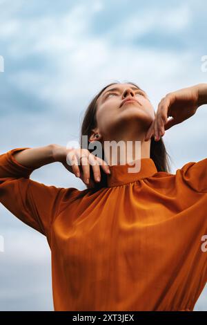 Dal basso la donna bruna contemplativa in un vivace abito arancione guarda verso l'alto, le sue mani toccano delicatamente il collo contro uno sfondo nuvoloso. Foto Stock