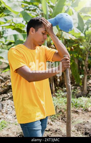 Un giovane vestito con una t-shirt gialla brillante e jeans si erge in un giardino illuminato dal sole, spazza il sudore dalla fronte e tiene in mano uno strumento da giardinaggio Foto Stock