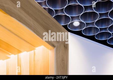 Un design del soffitto composto in modo creativo che combina pannelli in legno lucidato e tubi in metallo disposti simmetricamente, diffondendo un morbido bagliore ambientale da un embed Foto Stock