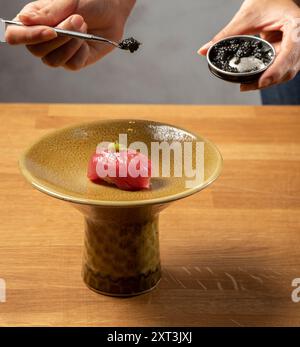 Un'immagine ravvicinata che mostra le mani usando delle pinzette per guarnire un pezzo di sushi con caviale, posto delicatamente su un piatto di ceramica ruvida Foto Stock