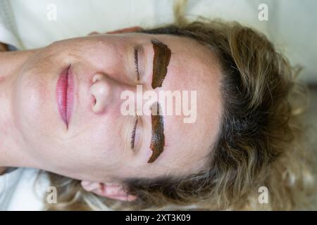 Vista ravvicinata di una donna che si stende con gli occhi chiusi, mostrando il processo di microblading sulle sopracciglia ricoperte di pasta sagomante scura. Foto Stock