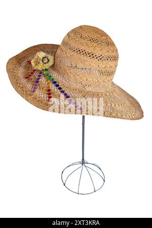 Cappello estivo in paglia con strass posizionato su un supporto isolato. L'elegante cappello a teglia larga ha un design intrecciato con decorazioni in fiori di paglia sul lato del bordo del cappello. Foto Stock