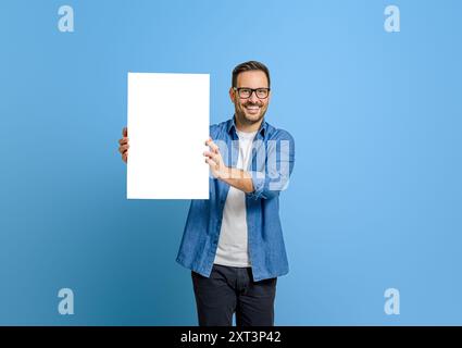 Ritratto del manager uomo felice che mostra un poster bianco vuoto per il marketing su sfondo blu Foto Stock