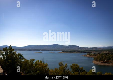 Il lago artificiale Gabriel y Galan di Caceres cattura un tranquillo paesaggio di acque blu, incorniciato da montagne lontane e alberi lussureggianti, offrendo un ambiente sereno Foto Stock