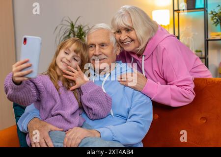 Nonno caucasico, nonna e piccola nipote che fanno videochiamate online su smartphone a casa. Ragazza con nonni che parla tramite webcam Internet, app chat seduta sul divano in soggiorno. Foto Stock