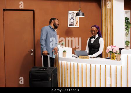 Receptionist afro-americana che utilizza un computer portatile mentre si trova dietro la reception e parla con gli ospiti dell'hotel. Un turista di colore che fa il check-in al resort. Vacanze in residence Foto Stock