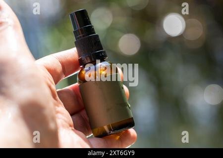 Primo piano di una mano che tiene in mano una piccola bottiglia spray in vetro marrone con sfondo naturale sfocato. Foto Stock