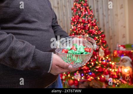 Una persona tiene in mano una ciotola di cristallo piena di cioccolatini davanti a un albero di Natale decorato adornato con ornamenti e luci rosse, che rappresenta gre Foto Stock