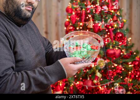 Una persona tiene in mano una ciotola di cristallo piena di cioccolatini davanti a un albero di Natale decorato adornato con ornamenti e luci rosse, che rappresenta gre Foto Stock