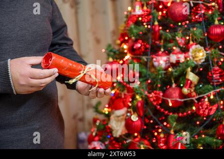 Una persona sta tenendo in mano un regalo festivo con un nastro verde davanti a un albero di Natale decorato in modo luminoso adornato con ornamenti e luci rosse Foto Stock