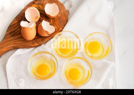 Uova incrinate in piccole ciotole di vetro: Uova crude incrinate in piccole ciotole di preparazione di vetro su un ripiano di cucina in marmo Foto Stock