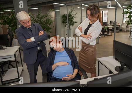 Un anziano uomo e una donna caucasica guardano disapprovatamente al loro collega incinta che dorme in ufficio. Foto Stock