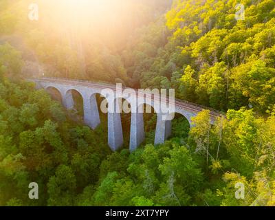 Il ponte ferroviario in pietra di Zampach si arrocca graziosamente su una lussureggiante vegetazione, illuminata dal caldo bagliore del sole che tramonta, mostrando l'armonia tra natura e ingegneria. Foto Stock