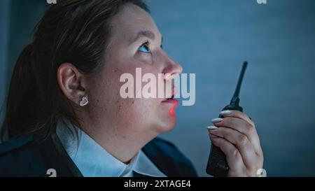 Primo piano di una donna che si occupa della sicurezza o di un operatore che parla in walkie talkie e guarda gli schermi seduti sul posto di lavoro nel centro di monitoraggio durante il turno di notte. Sistema di sorveglianza e CCTV. Verticale. Foto Stock