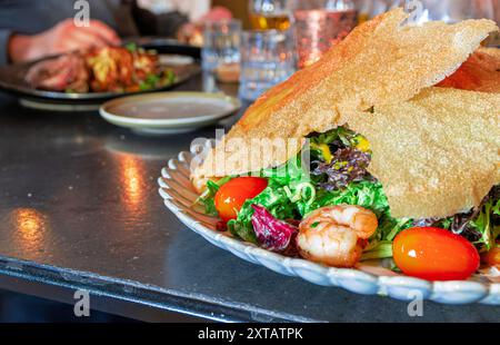 Un piatto di cibo con gamberi, pomodori e una tortilla croccante. Il piatto si trova su un tavolo con altri piatti e bicchieri Foto Stock