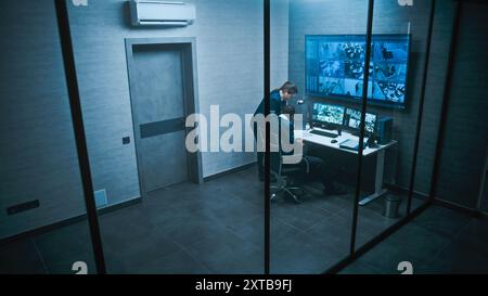 L'operatore afroamericano CCTV lavora su computer guardando le telecamere di sicurezza in ufficio. Riprese di sorveglianza su più monitor e grande schermo digitale. Una lavoratrice donna entra e parla con una collega. Foto Stock