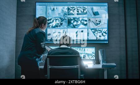 Una donna della sicurezza parla con una collega afroamericana, usa un tablet. Gli operatori di sorveglianza multietnica controllano le telecamere di sicurezza sui monitor dei computer e sul grande schermo digitale con filmati CCTV. Foto Stock