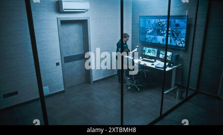 L'operatore afroamericano CCTV lavora su computer guardando le telecamere di sicurezza in ufficio. Riprese di sorveglianza su più monitor e grande schermo digitale. Una lavoratrice donna entra e parla con una collega. Foto Stock