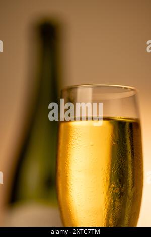 Primo piano di un flute di champagne ghiacciato pieno di champagne frizzante. Una bottiglia di champagne verde è sfocata sulla destra, con una sfumatura d'oro verticale Foto Stock