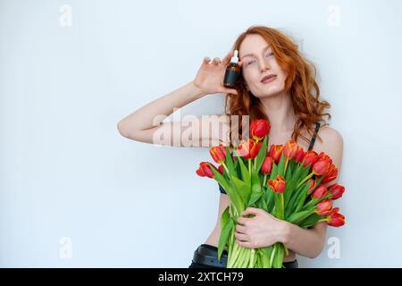 Una donna tiene in mano il profumo della bottiglia e un bouquet di tulipani rossi. indossa un reggiseno nero e una camicia nera Foto Stock