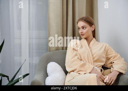 Una giovane donna si rilassa in un morbido accappatoio, seduto su un comodo divano vicino a una finestra. Foto Stock