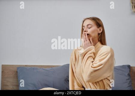 Una giovane donna in vestaglia si siede sul suo letto e sbadiglia. Foto Stock