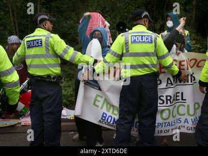 Leicester, Leicestershire, Regno Unito. 14 agosto 2024. I manifestanti si trovano di fronte alla fabbrica gridando e tenendo striscioni trattenuti da un cordone di polizia durante la dimostrazione. I manifestanti si riuniscono fuori dai cancelli della fabbrica Elbit System a Leicester. Gli operatori sanitari per la Palestina si sono Uniti alla manifestazione molti di loro nei loro scrub per protestare mentre i lavoratori venivano a lavorare presso la compagnia israeliana di armi. I manifestanti protestano contro le armi fabbricate in questa fabbrica che ritengono vengano successivamente utilizzate per opprimere i palestinesi a Gaza e altrove. Il 85% della flotta israeliana di droni è fornito da El Foto Stock